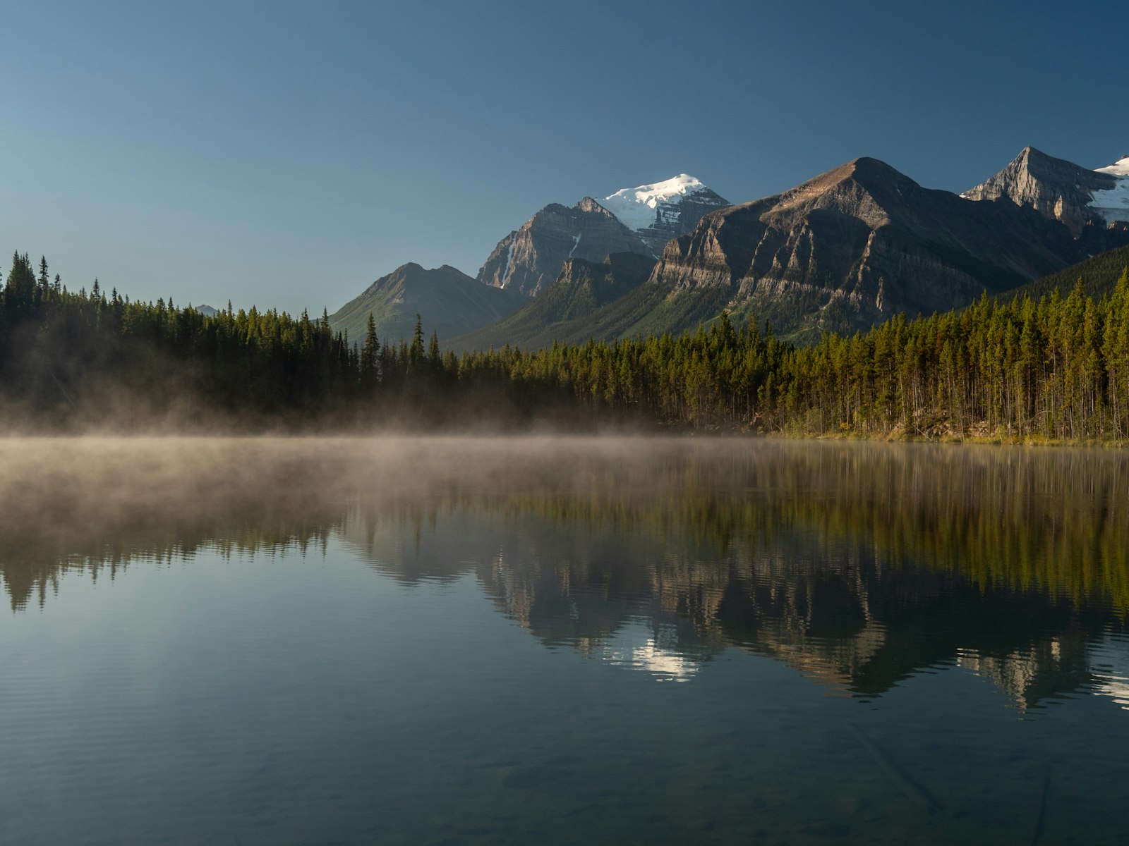 Lago parado ao amanhecer refletindo montanhas e névoa — água quieta é o que deixa ver o fundo